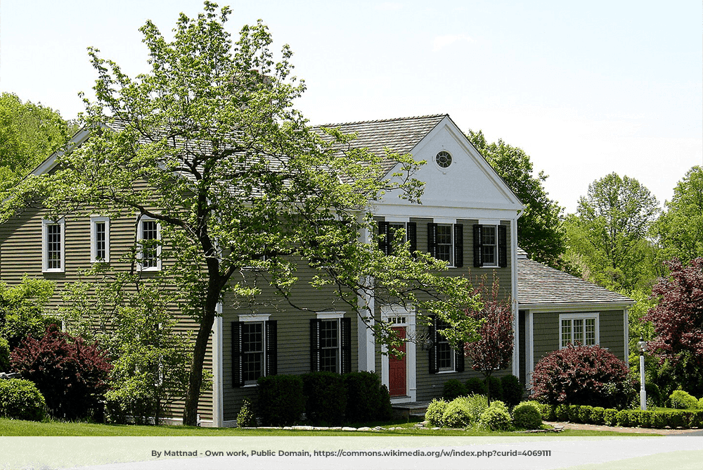A large two-story colonial-style house with olive green siding, white trim, and black shutters, partially obscured by leafy trees and surrounded by well-maintained bushes and landscaping.