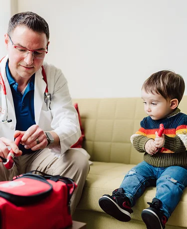 A doctor in a white coat sits on a sofa, looking at a small child's knee, with the child sitting next to him.