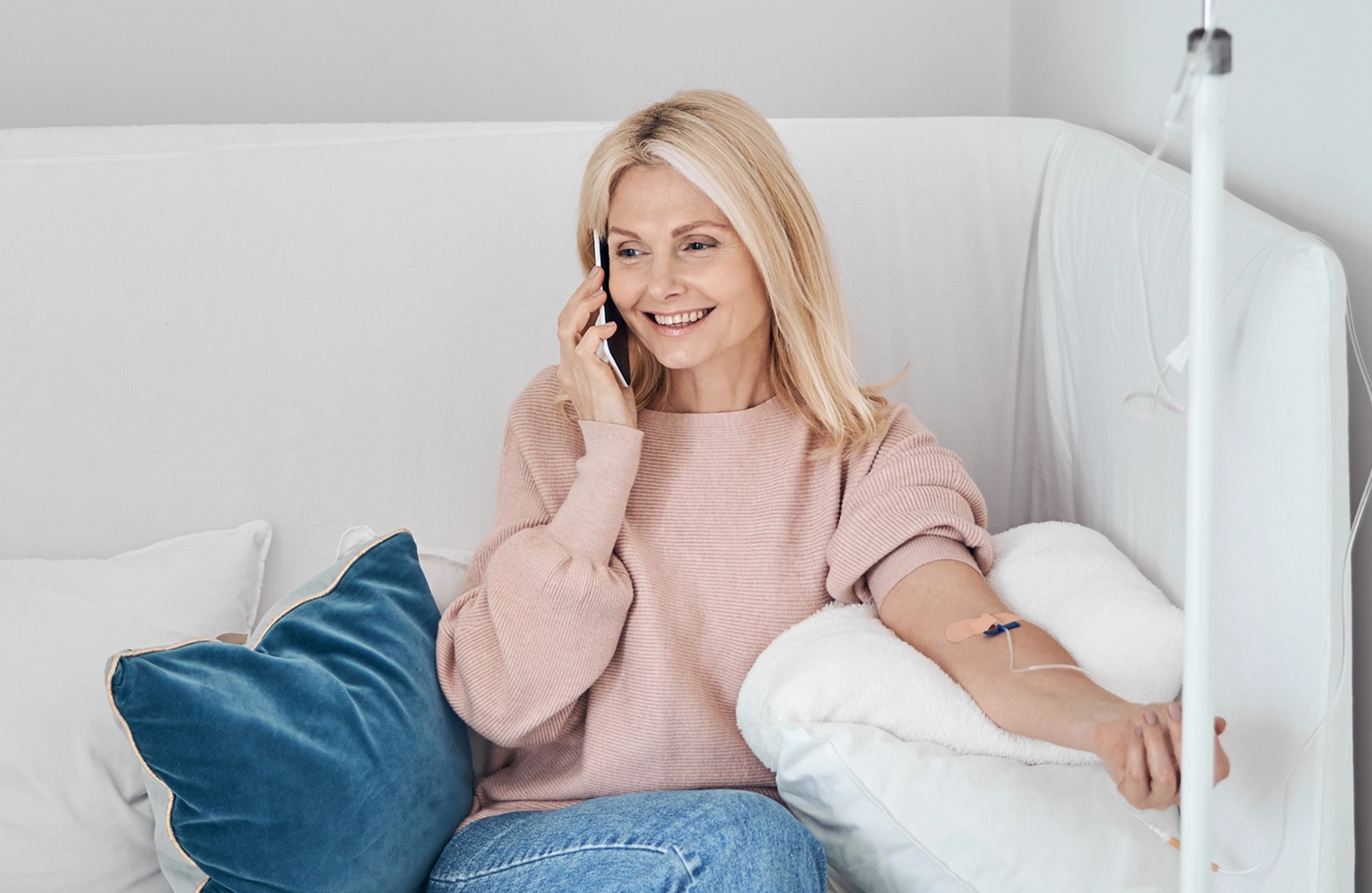 A healthcare professional using a stethoscope to examine a smiling female patient in her living room, with a clinical IV hydration setup positioned nearby.