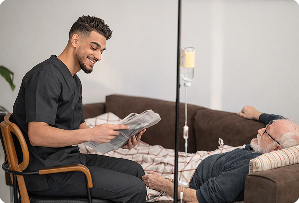 A male nurse in black clinical scrubs smiling and interacting with a senior male patient resting on a sofa during an at-home IV therapy session.