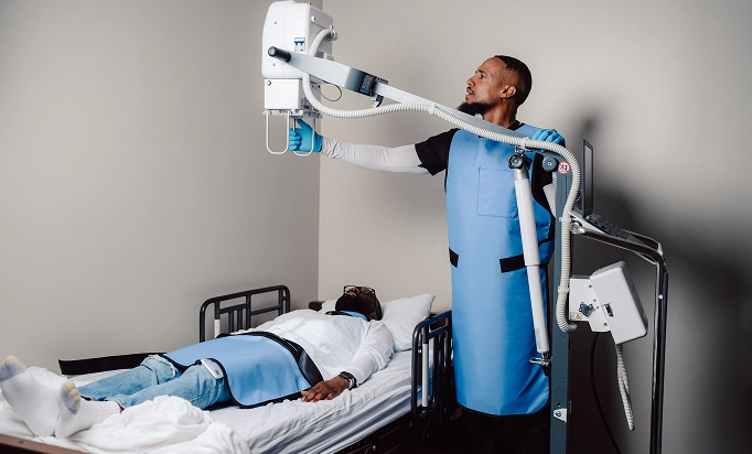A Life Mobile Care X-ray technologist conducting a bedside imaging procedure for a patient in a medical facility, both wearing protective aprons.